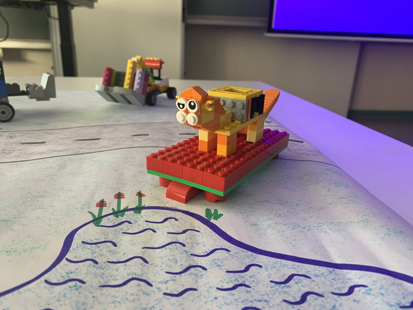 Ein kleiner Löwe als Lego-Modell auf einer gezeichneten Unterlage, die eine Siese und einen Teich darsellt